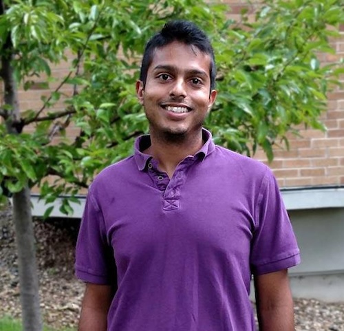 Man in a purple shMan in a purple shirt smiling for the camera in front of a brick building with a tree growing beside it