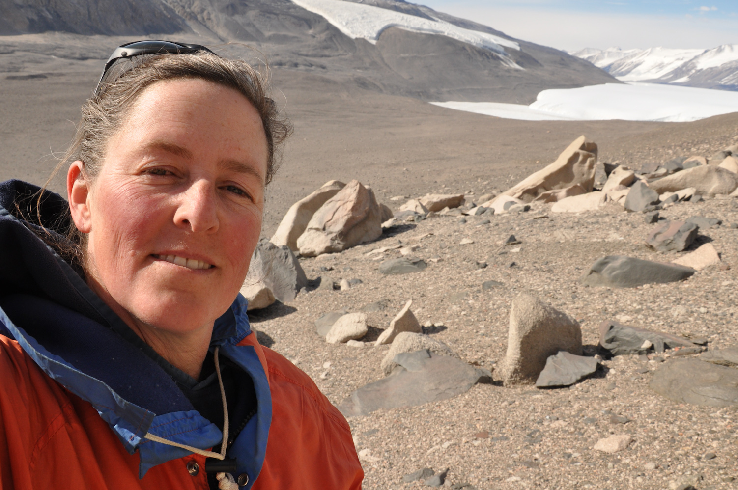 selfie of a woman in a red jacket standing on a barren landscape with snow topped mountains in the background