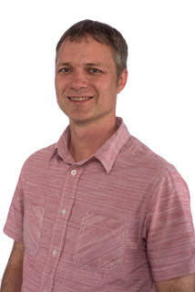 photo of a man in a light red button up shirt smiling at the camera in front of a white backdrop