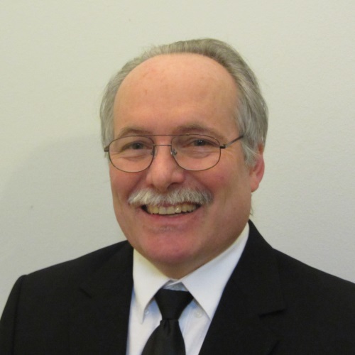 professional photo of a man in a black suit jacket, white shirt, and tie smiling for the camera in front of an off white backdrop